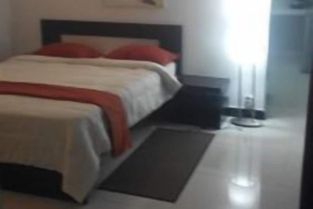 A bedroom with a bed, a bedside table, a nightstand, a window with curtains, and a wall-mounted light fixture.
