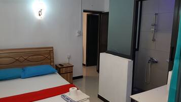 A hotel room with a bed, a bedside table, a remote control, a roll of toilet paper, and a shower area.