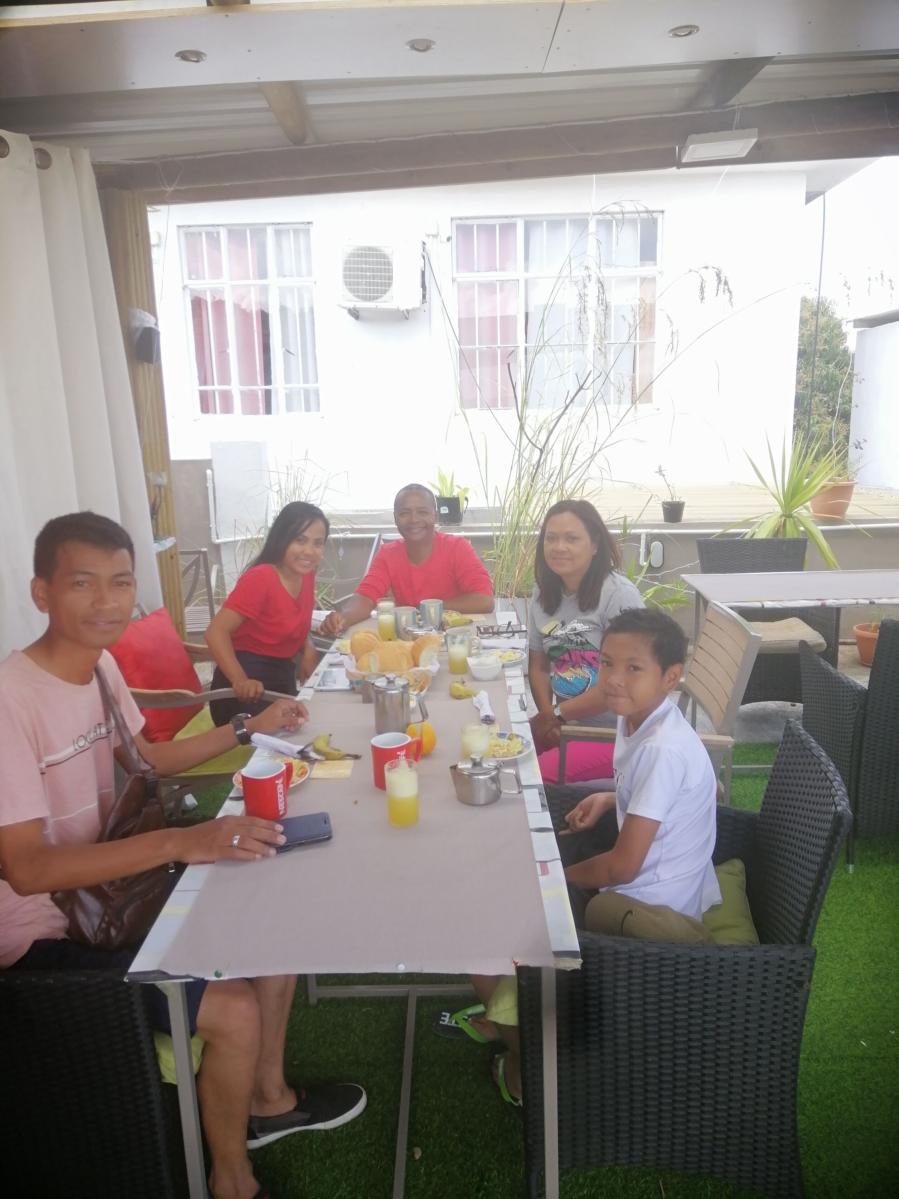 A group of people seated around a table with food and drinks, outdoors with a building in the background.