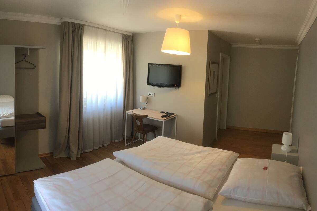Habitación de hotel con dos camas, un escritorio con silla, televisor montado en la pared y una ventana con cortinas.