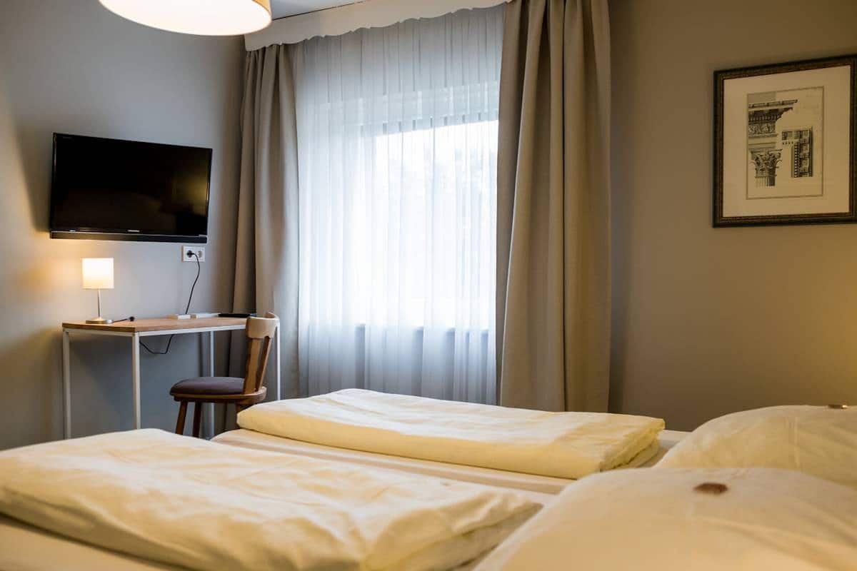 Habitación de hotel con dos camas individuales, un escritorio con silla, un televisor montado en la pared y una ventana con cortinas de gasa.