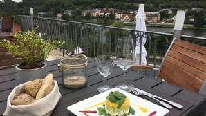 Une table à manger avec un repas, un bocal en verre contenant une bougie, et une vue sur une rivière et des maisons en arrière-plan.