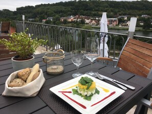 Een eettafel met een maaltijd, een glazen pot met een kaars, en een rivier en huizen op de achtergrond.