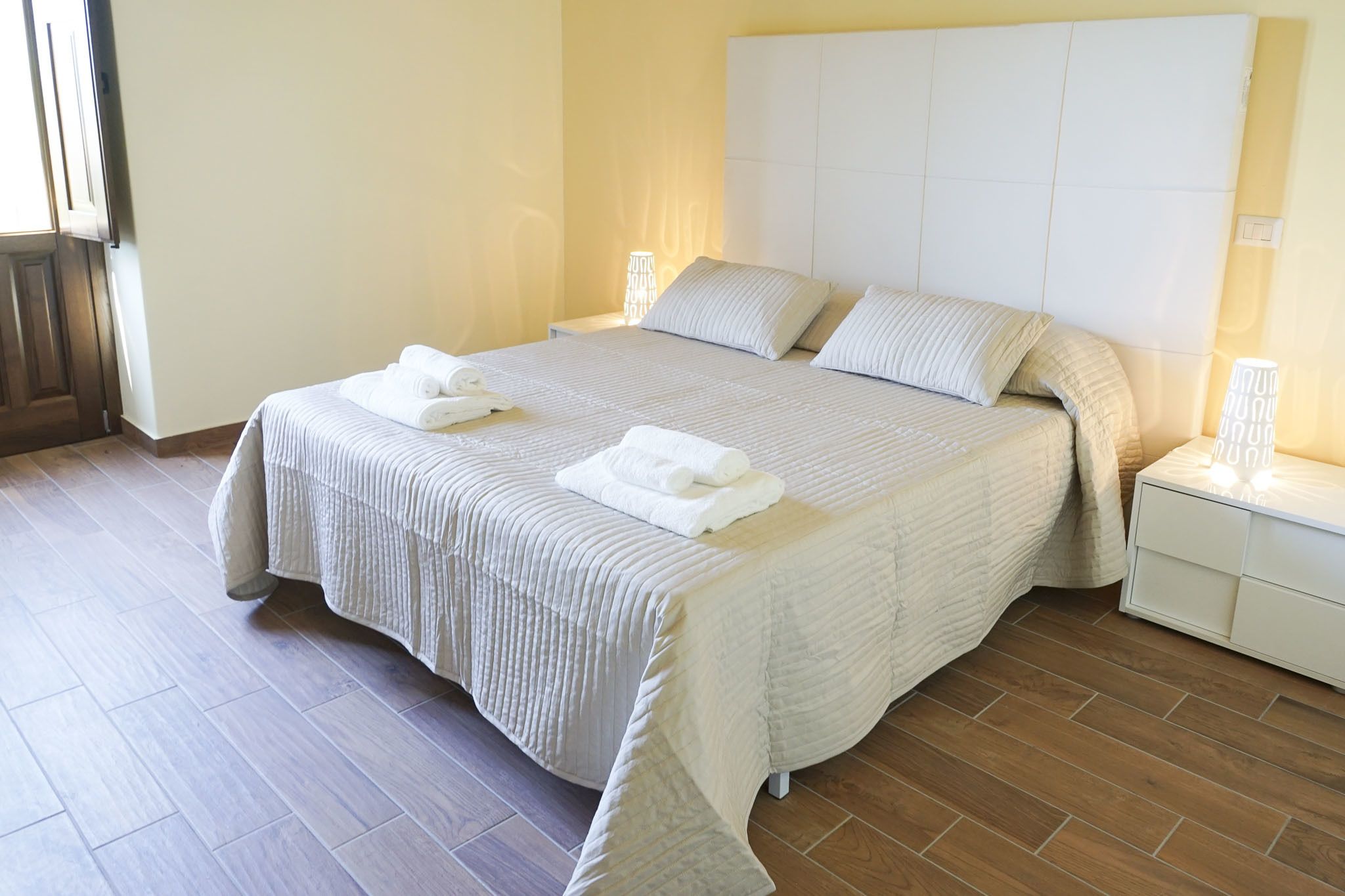 Una cama bien hecha con sábanas y almohadas blancas, una mesita de noche con una lámpara y suelo de madera.