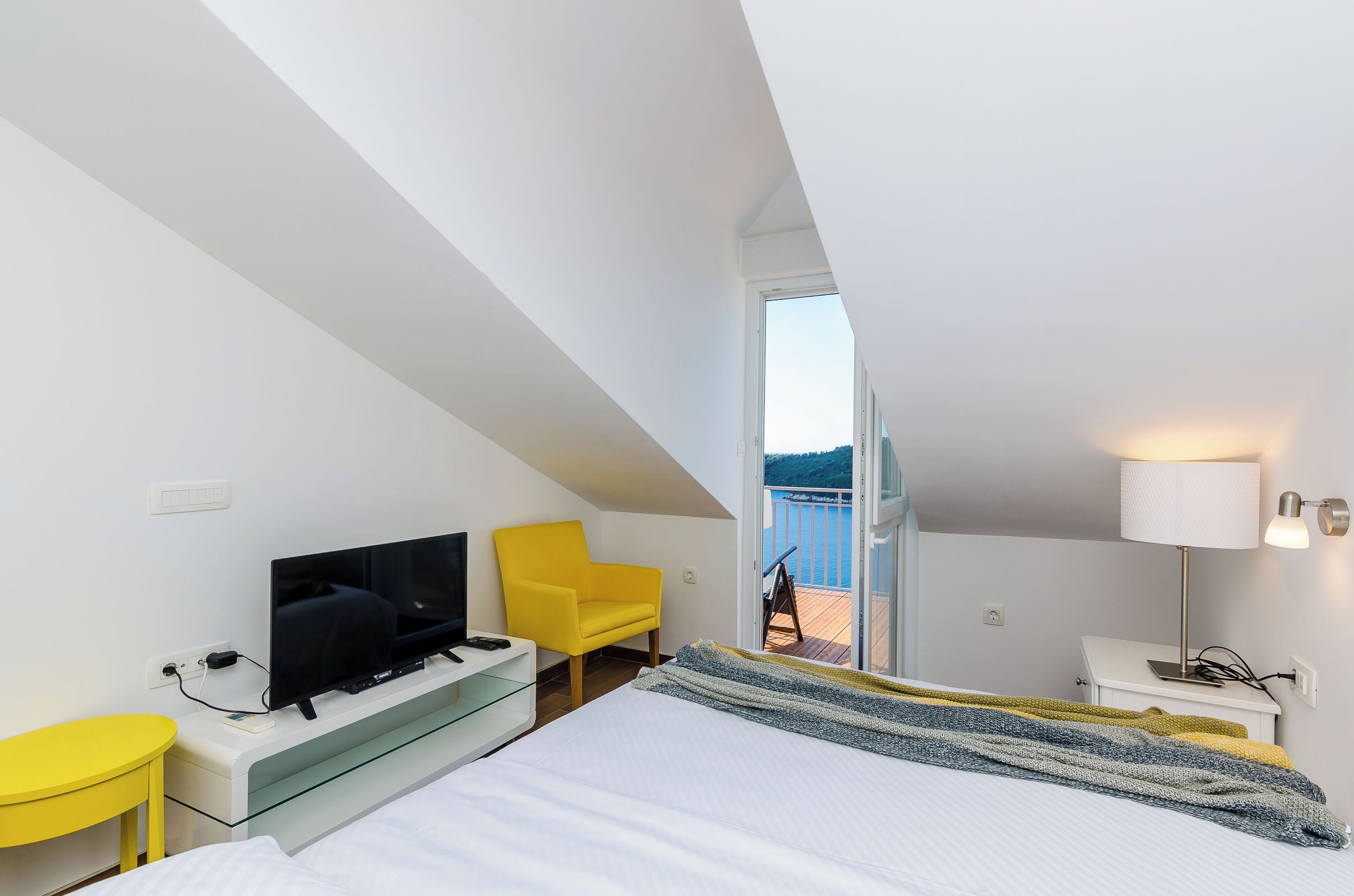A modern hotel room with a bed, a TV, a yellow chair, and a balcony with a view.