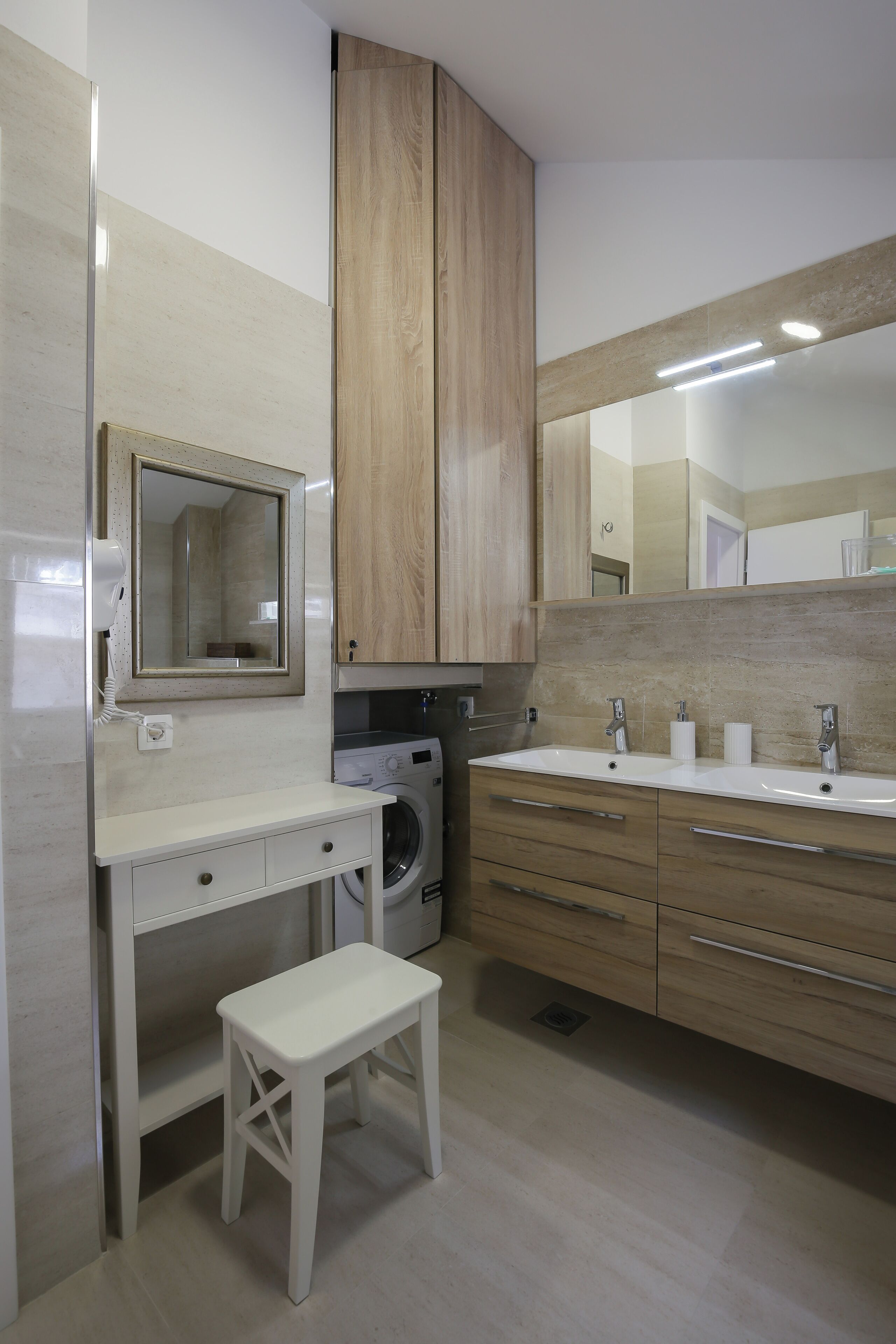 Een moderne badkamer met een ingebouwde wasmachine, een dubbele wastafel en een klein krukje.
