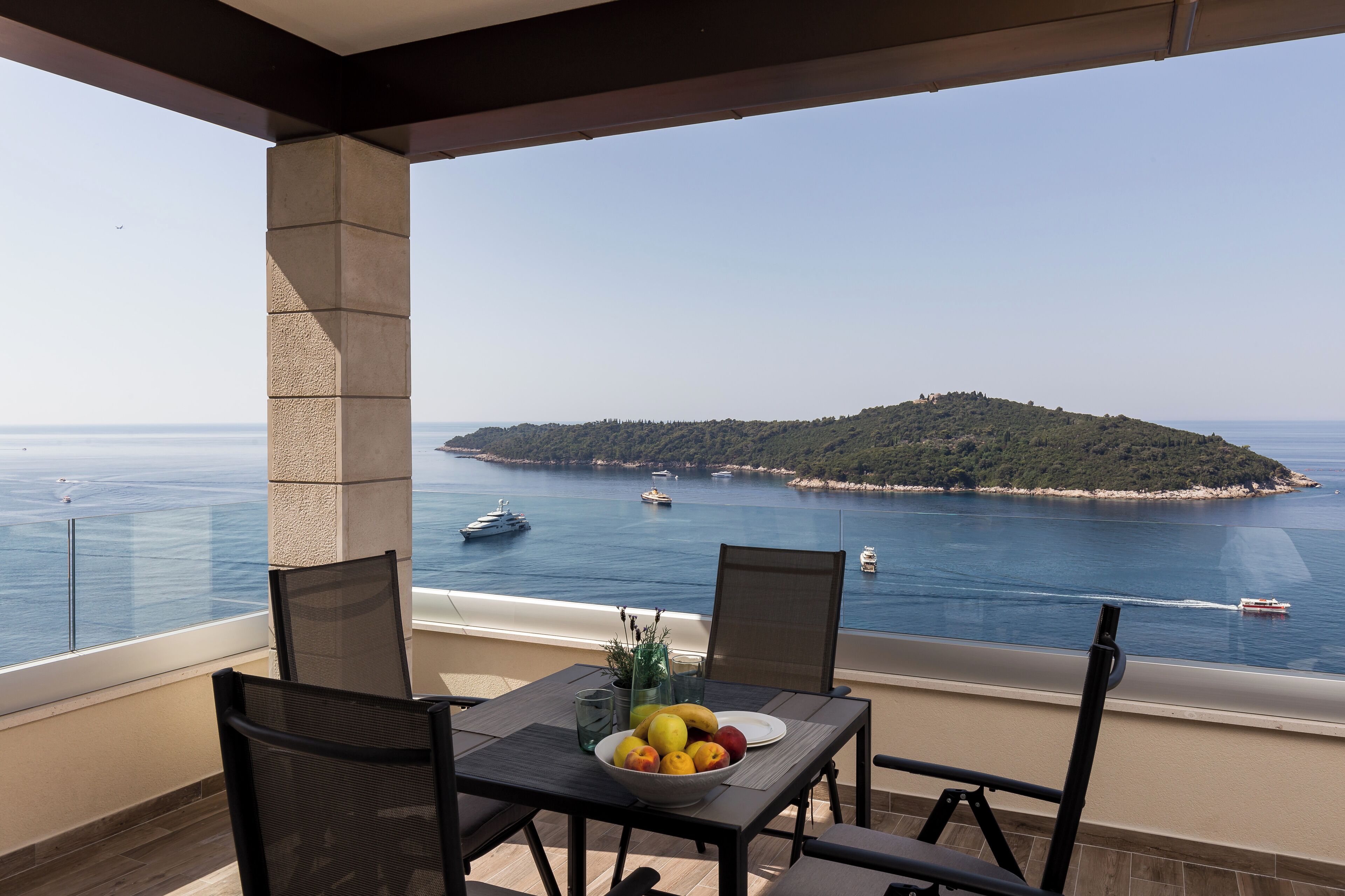 Balcón con mesa lista para comer, con vista al mar y barcos, e isla pequeña.