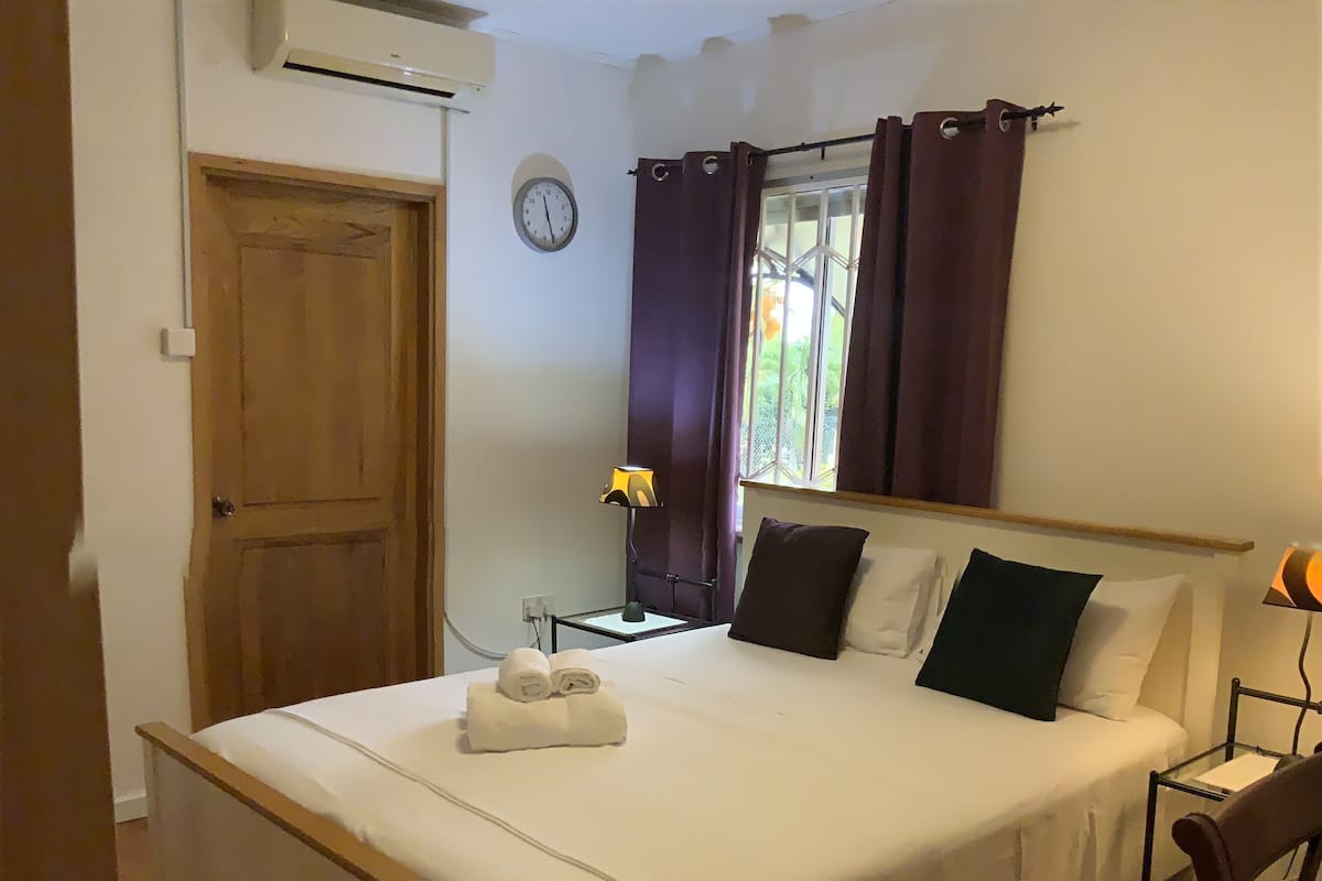 Una cama bien hecha con una toalla, una mesita de noche con una lámpara, un reloj en la pared y una ventana con cortinas.