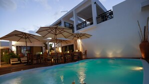 Uma área à beira da piscina com guarda-sóis, uma mesa de jantar e cadeiras.