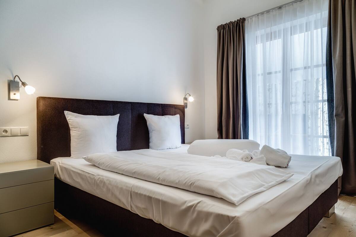 A neatly made bed with white linens, a dark headboard, and two wall-mounted reading lights.