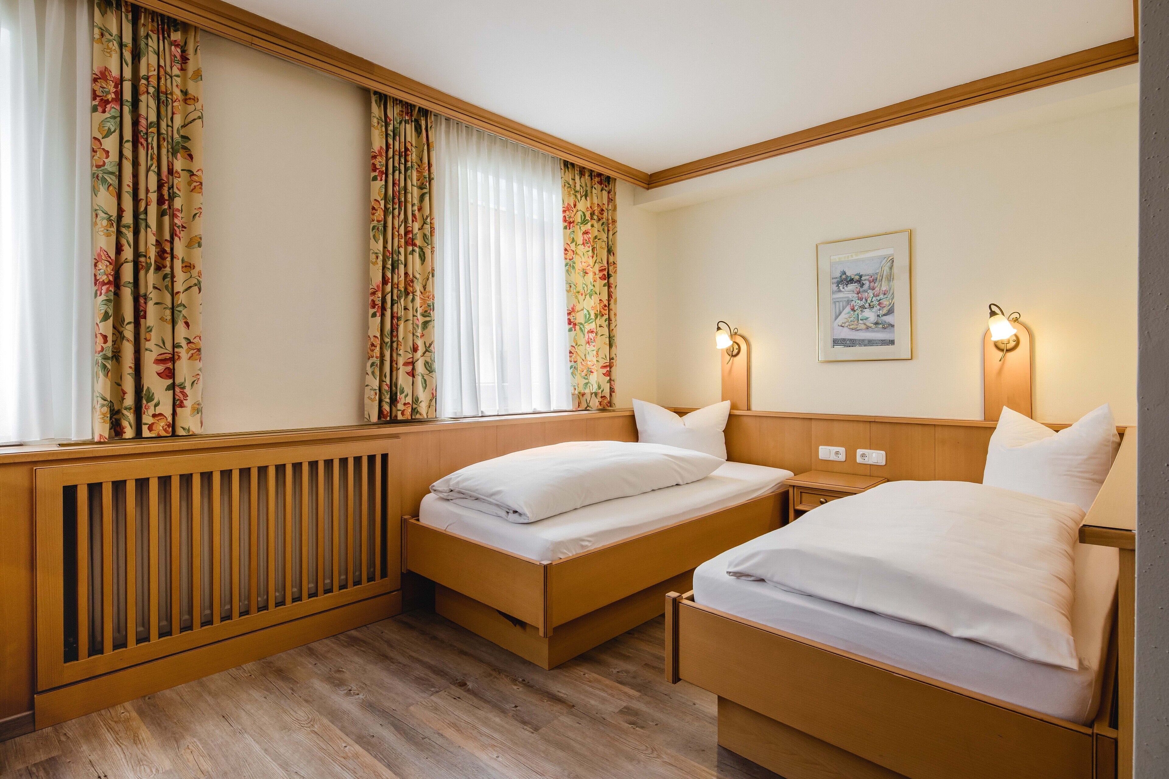 A hotel room with two single beds, wooden headboards, a radiator, floral curtains, a framed picture on the wall, and bedside lamps.