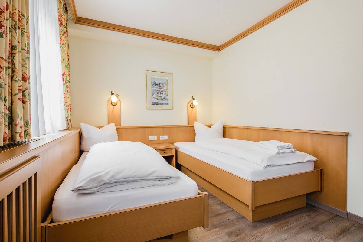 A hotel room with two wooden beds, a nightstand, a wall-mounted lamp, and a framed picture on the wall.
