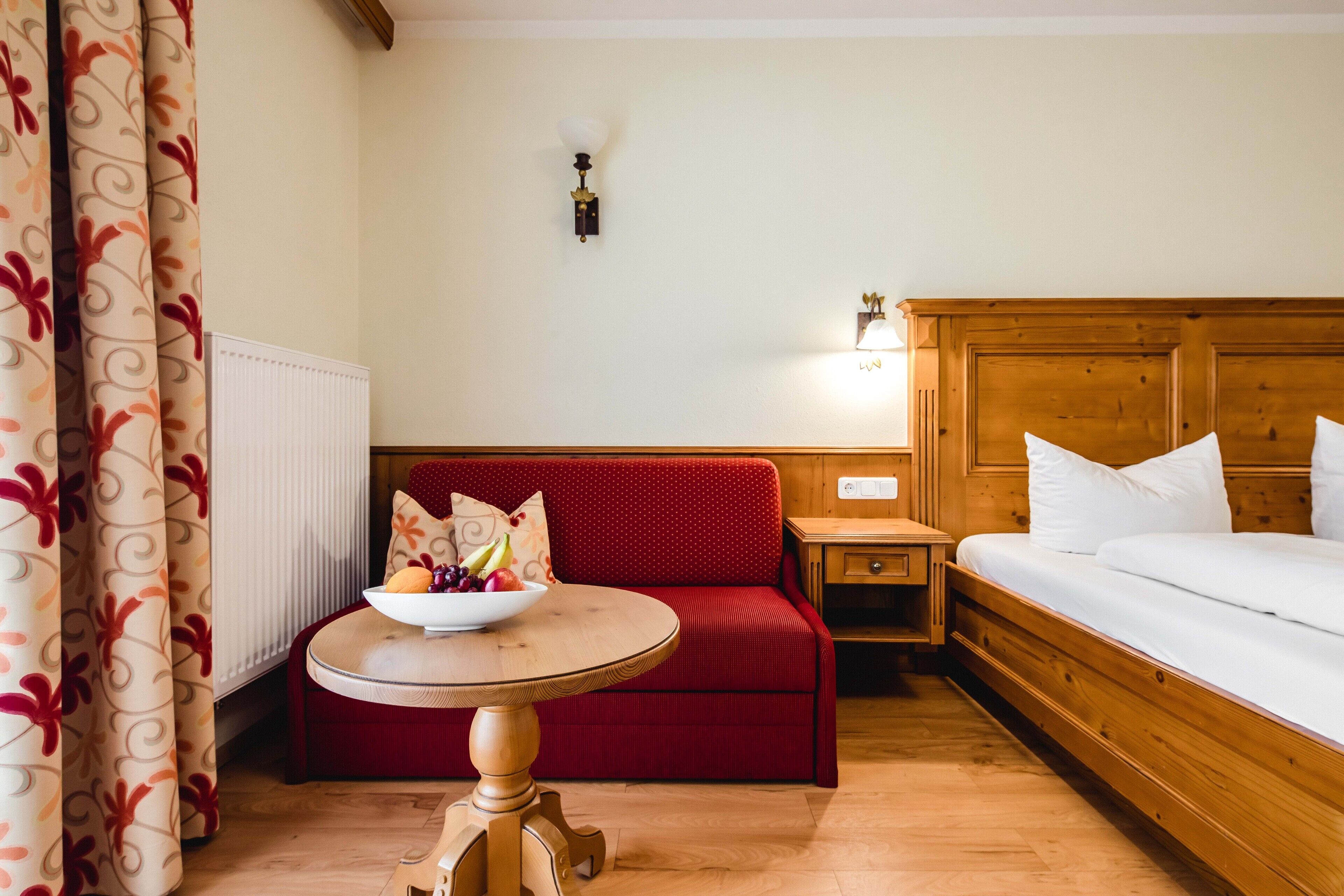 A hotel room with a wooden bed, a red sofa, a small table with a fruit bowl, a nightstand, and a wall-mounted light fixture.