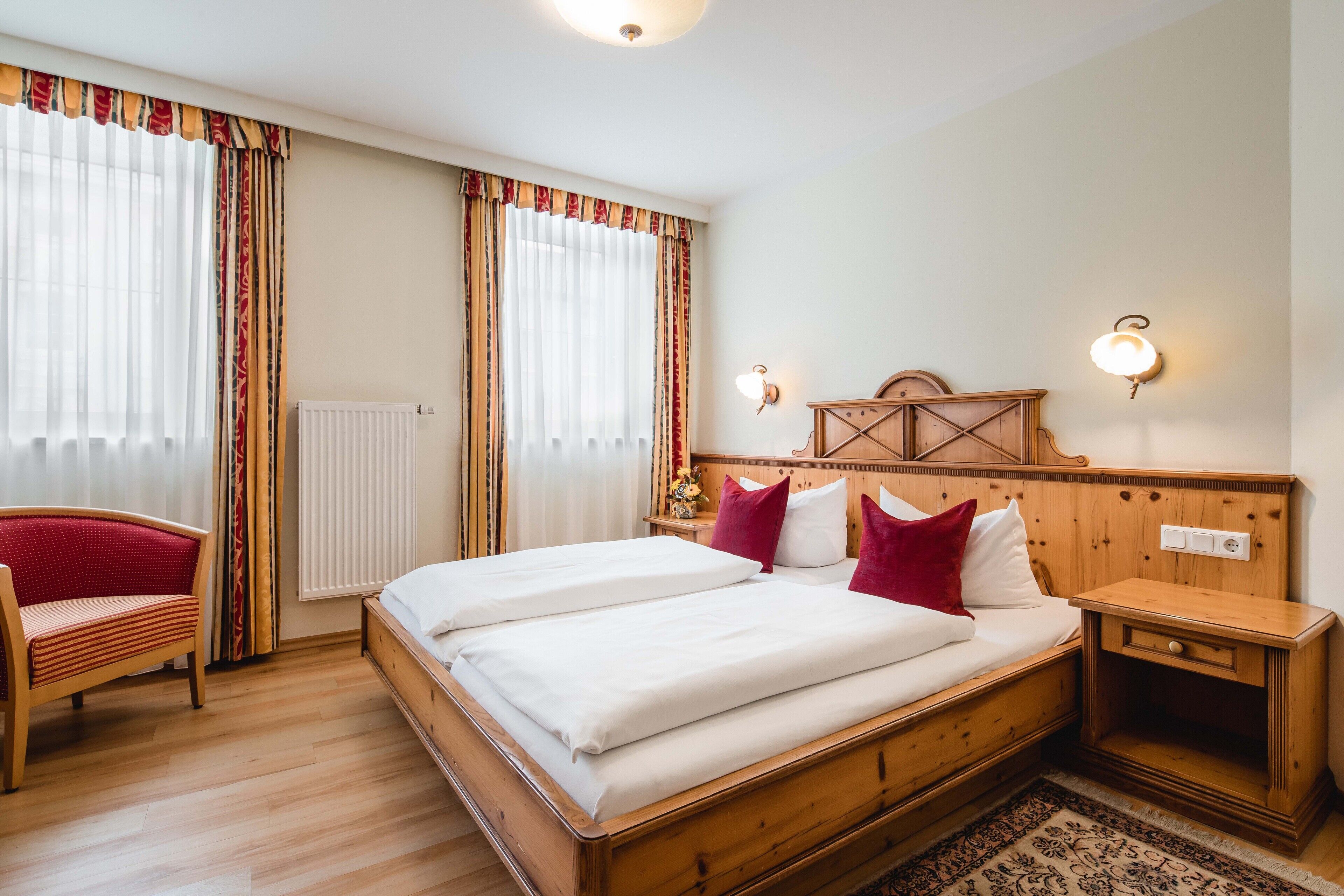 Una cama doble con ropa de cama blanca, cabecera de madera, dos mesitas de noche con lámparas, una silla roja y una ventana con cortinas.