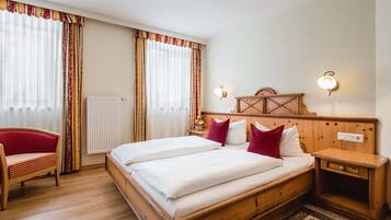Una cama doble con ropa de cama blanca, cabecera de madera, dos mesitas de noche con lámparas, una silla roja y una ventana con cortinas.