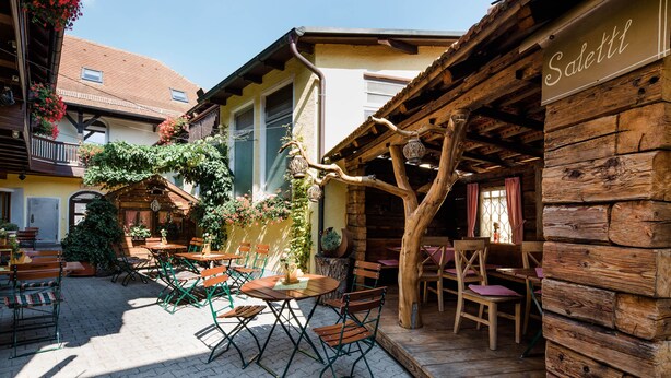 An outdoor café area with wooden tables and chairs, surrounded by buildings with a rustic architectural style. - Hotel bayrischer Löwe (Osterhofen)