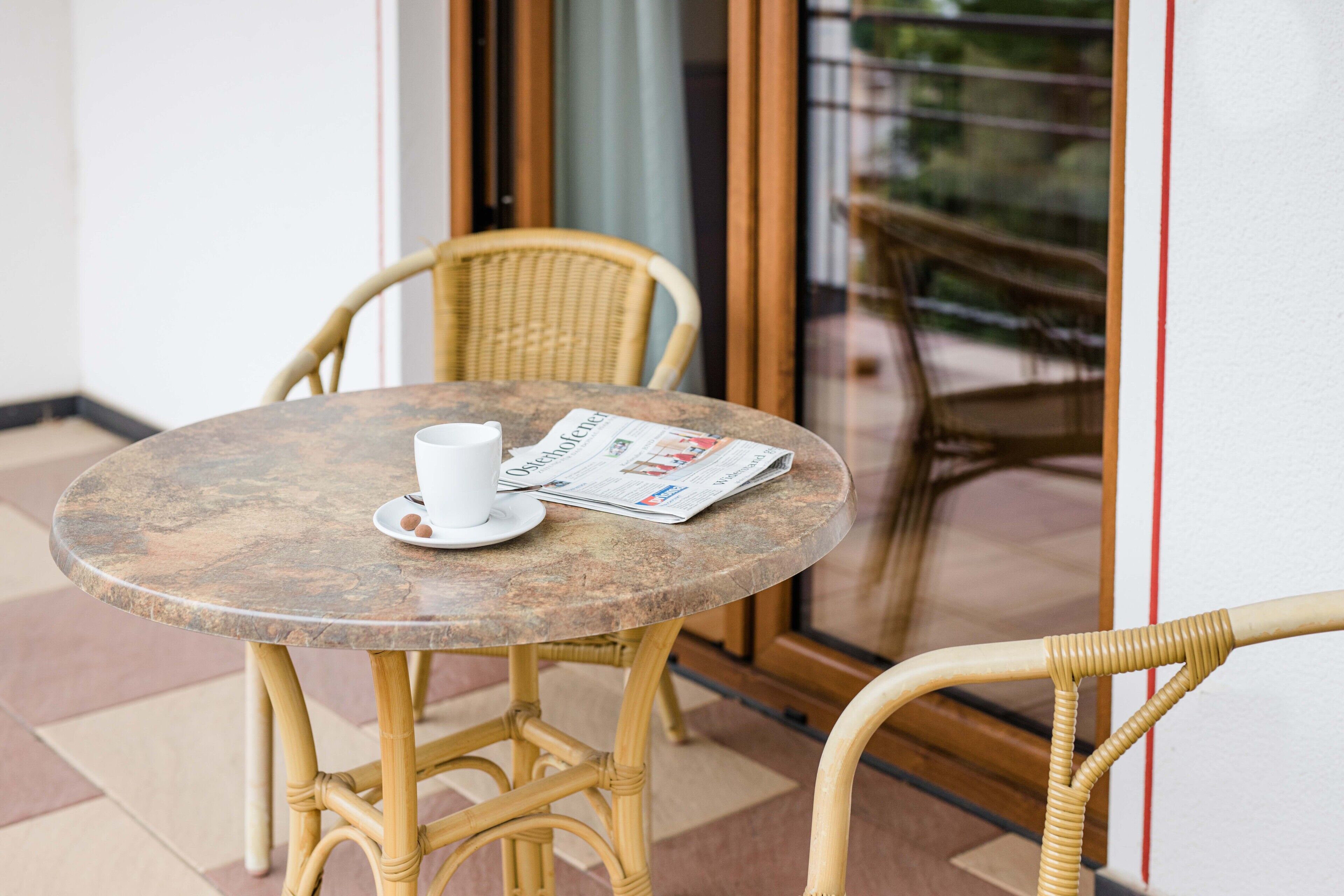 Una mesa redonda con una taza y un periódico, sillas de mimbre y un ventanal de vidrio que da a un espacio al aire libre.