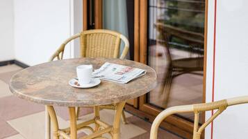 Una mesa redonda con una taza y un periódico, sillas de mimbre y un ventanal de vidrio que da a un espacio al aire libre.