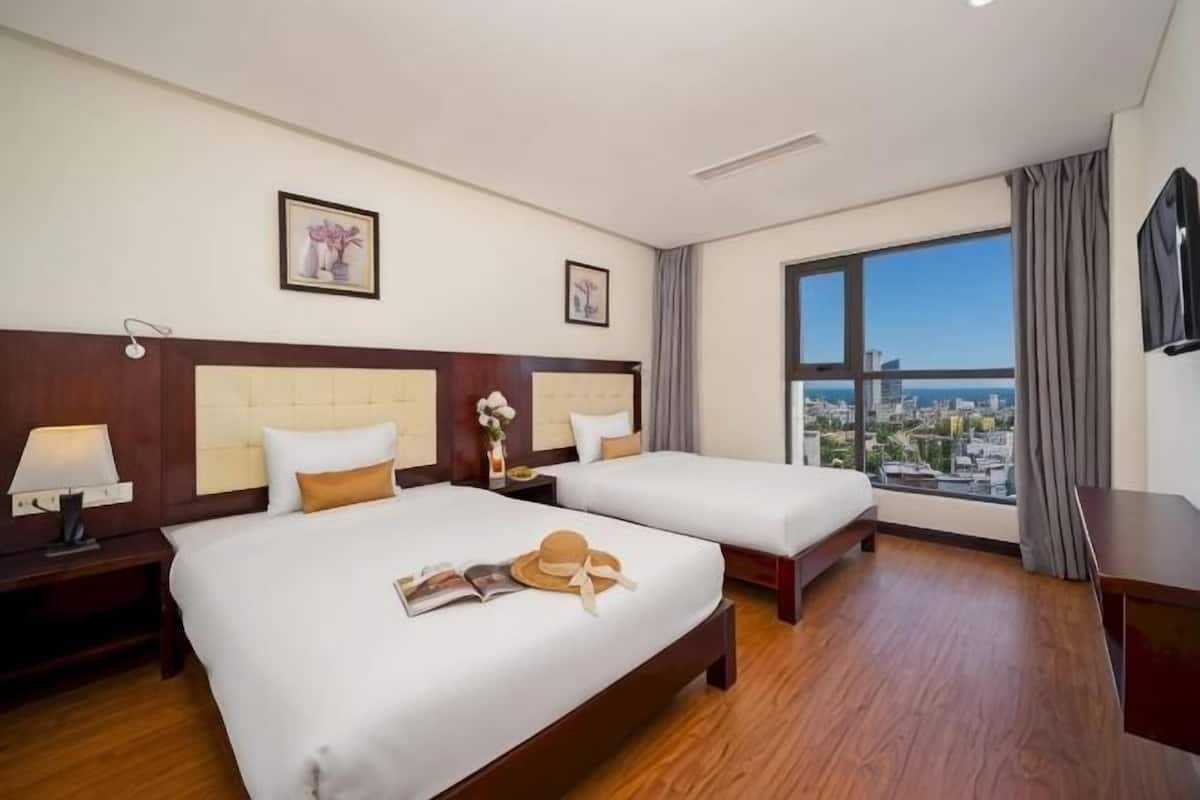 Una camera d'albergo con due letti, un comodino con una lampada, una finestra vista città e un quadro appeso al muro.