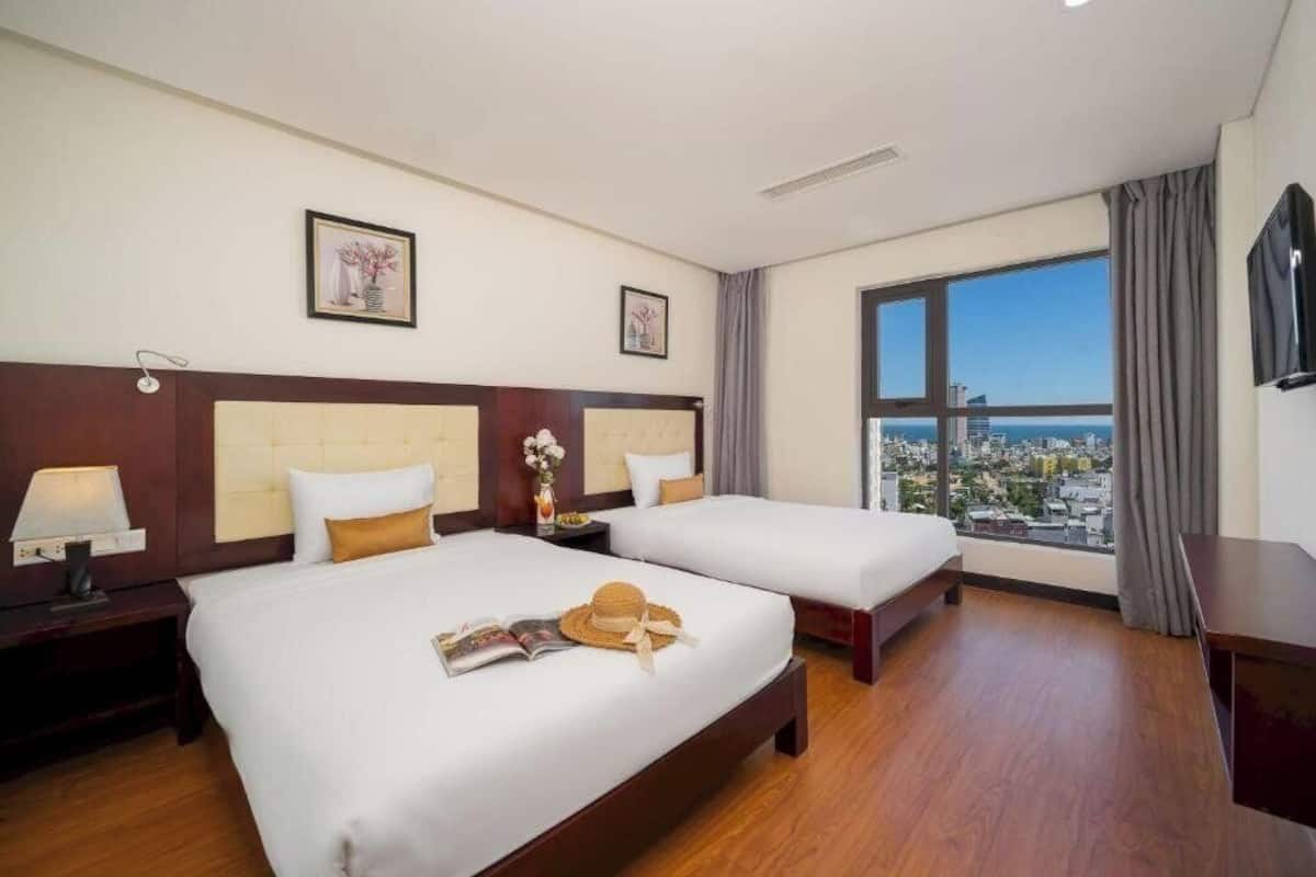 Una camera d'albergo con due letti, un'ampia finestra vista città e una televisione a schermo piatto appesa al muro.