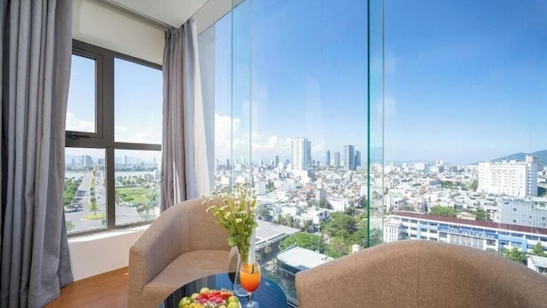 A hotel room with a large window offering a city view, a dining table with fruit and a drink, and two armchairs. - Gemma Hotel & Apartment Near Dragon Bridge  (Da Nang)