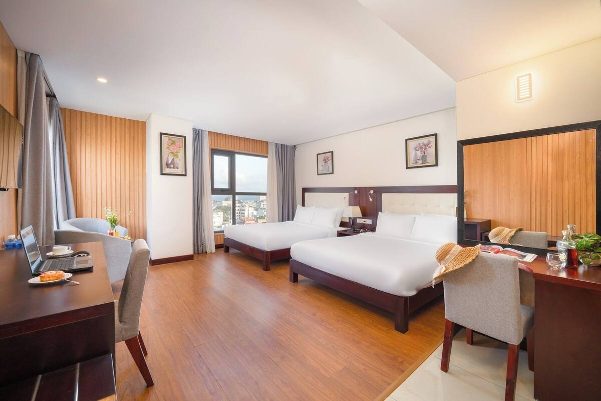 Una camera d'albergo con due letti, una scrivania con un portatile, una sedia, un tavolo da pranzo e vista sulla città.