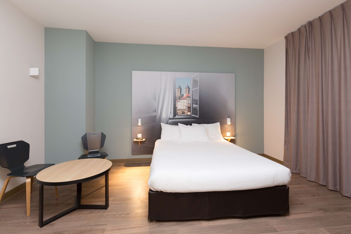 A modern hotel room with a large bed, a desk, and a chair. There is a painting on the wall and a view of buildings outside the window.