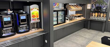 A modern café with self-service coffee machines, refrigerated display cases, and a variety of food items.