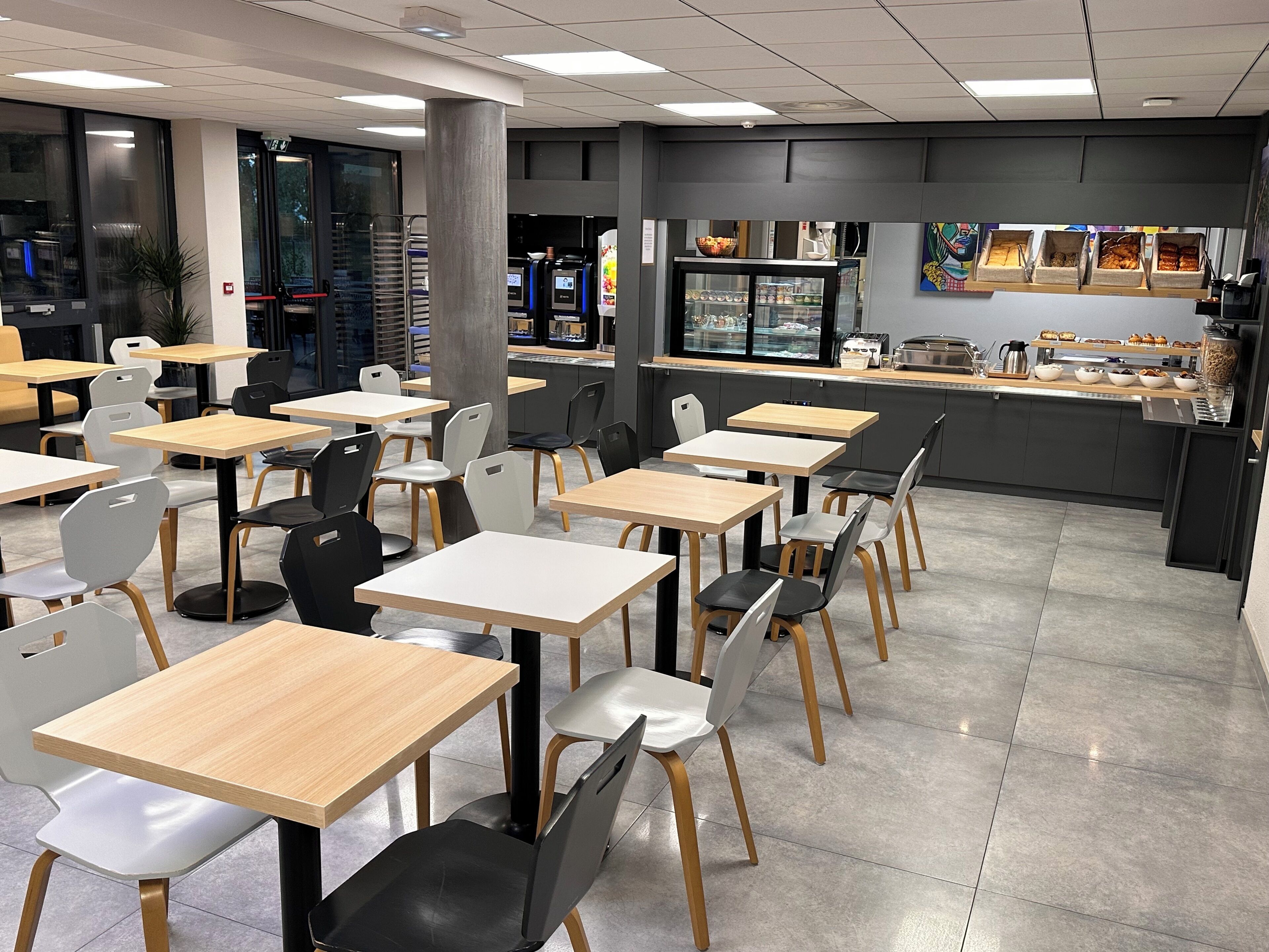 A modern cafe with multiple tables and chairs, a counter with food display, and a large window with a view.