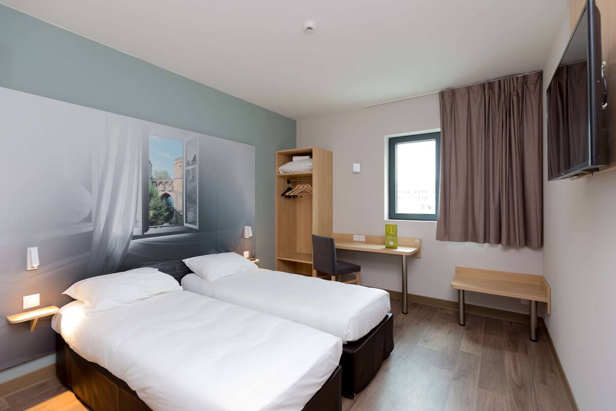 A hotel room with two beds, a desk, a chair, a TV, and a window with curtains.