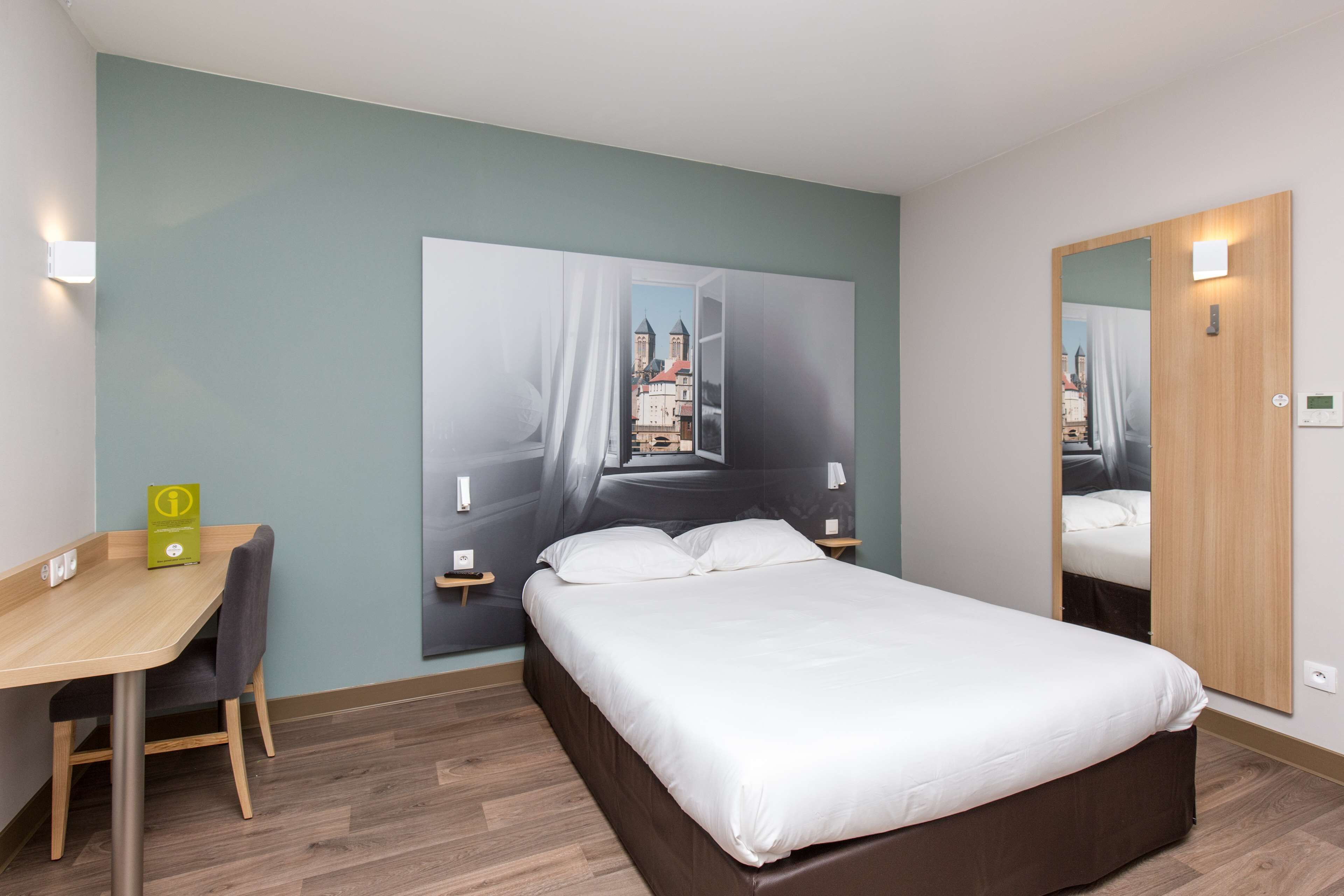 Ein Hotelzimmer mit Bett, Schreibtisch und Stuhl. Ein großes Fenster bietet Blick auf Gebäude.