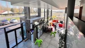 Un hall d’hôtel moderne avec de grandes fenêtres, un espace salon doté de meubles rouges et orange, et une vue sur les voitures garées à l’extérieur.