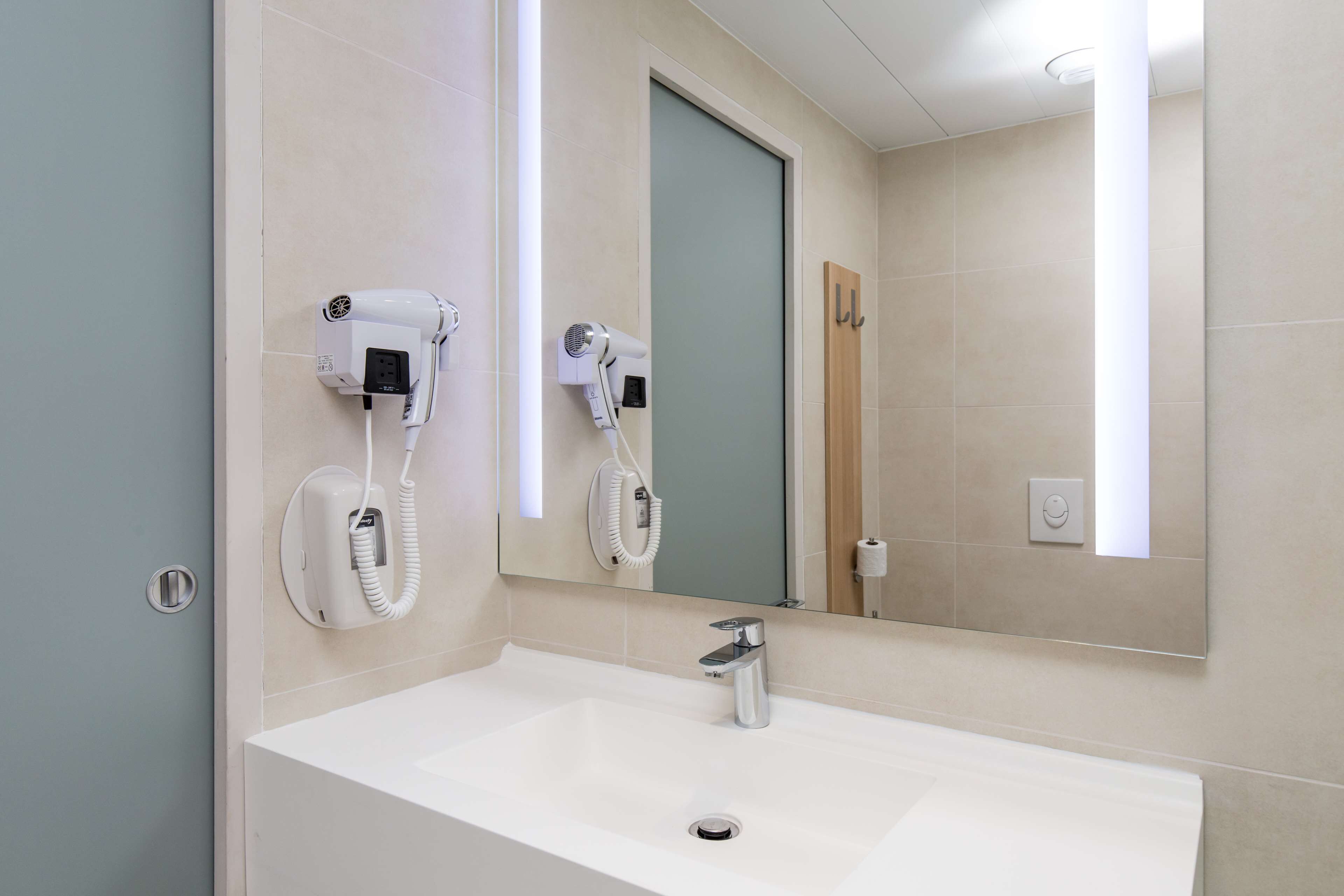 Un baño con un lavamanos blanco, dos secadores de pelo sujetos a la pared, un espejo y una puerta.