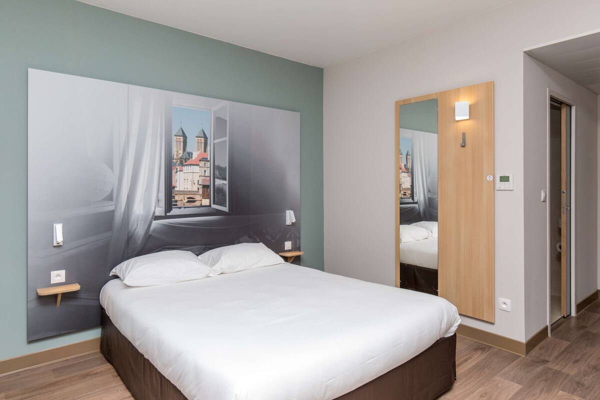 A modern hotel room with a large bed, a nightstand, a wall-mounted light, and a mirror.