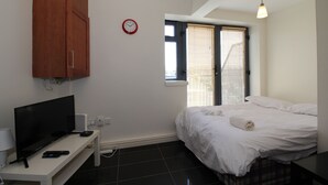A bedroom with a bed, a TV on a stand, a clock, and a window with blinds.