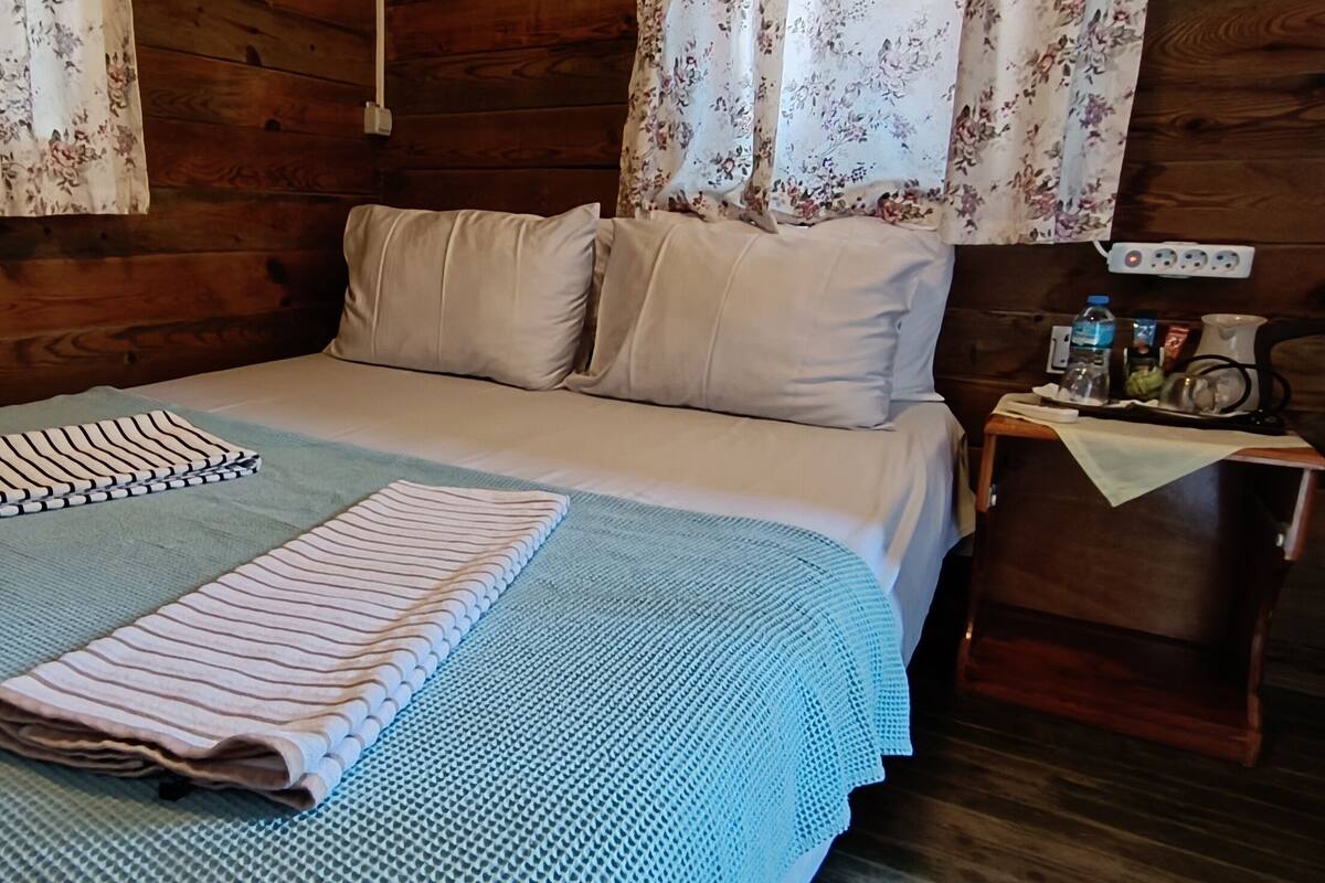 Ein ordentlich bezogenes Bett mit einer gestreiften Decke und einem gefalteten Handtuch, ein Nachttisch aus Holz mit einer Wasserflasche und einer Tasse sowie blumige Vorhänge an einer Holzwand.