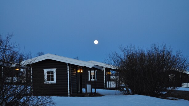 A wooden cabin with a snow-covered ground, a visible moon in the sky, and bare trees around. - Tana Familiecamping (Tana)