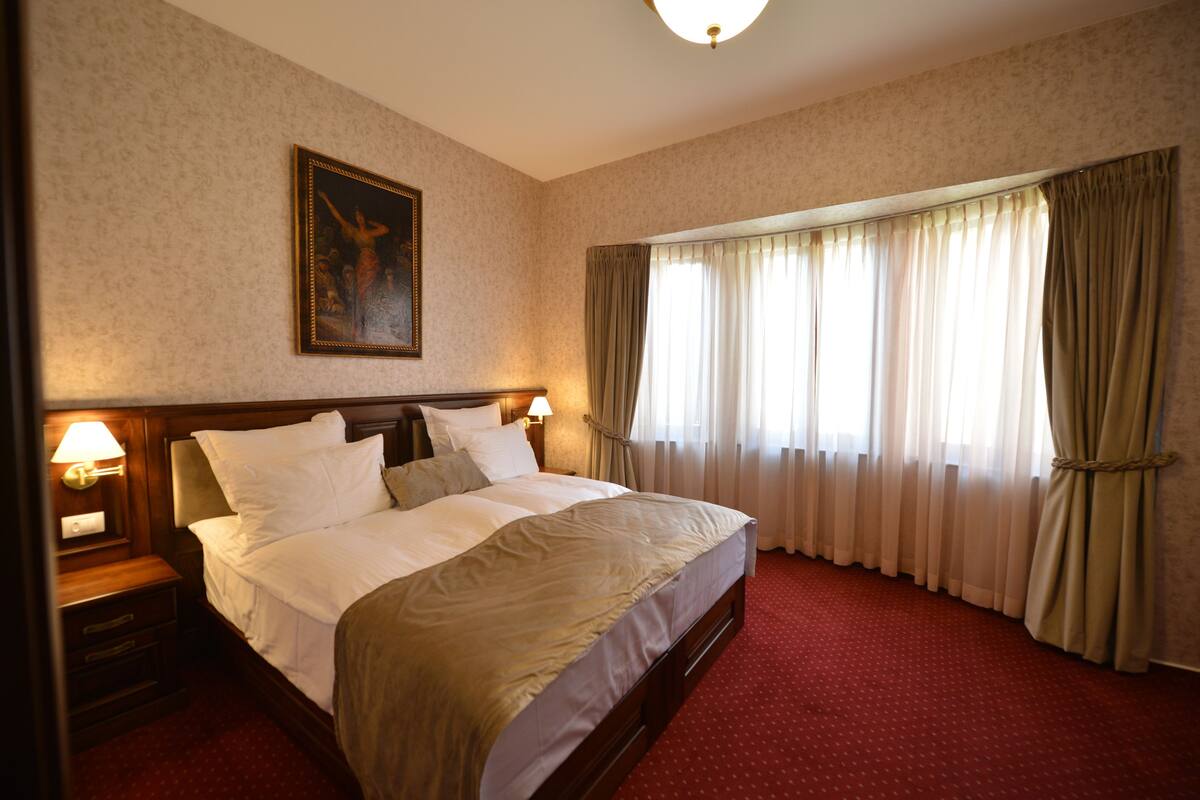A hotel room with a large bed, two bedside tables with lamps, a painting on the wall, and a window with curtains.