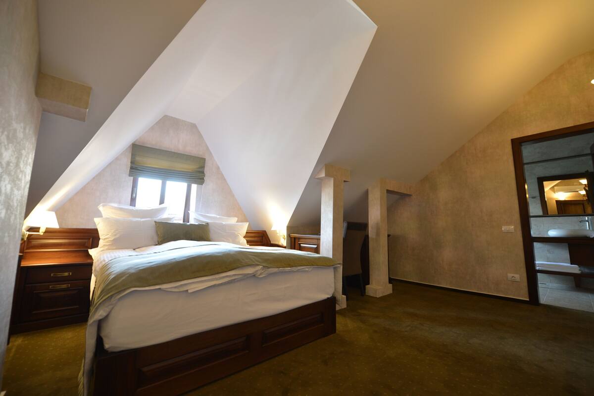 A bedroom with a sloped ceiling, a bed, bedside tables, and a bathroom visible through an open door.