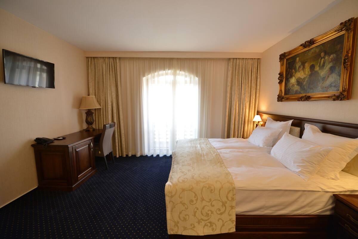 A hotel room with a large bed, a desk with a lamp, a TV, and a painting on the wall.
