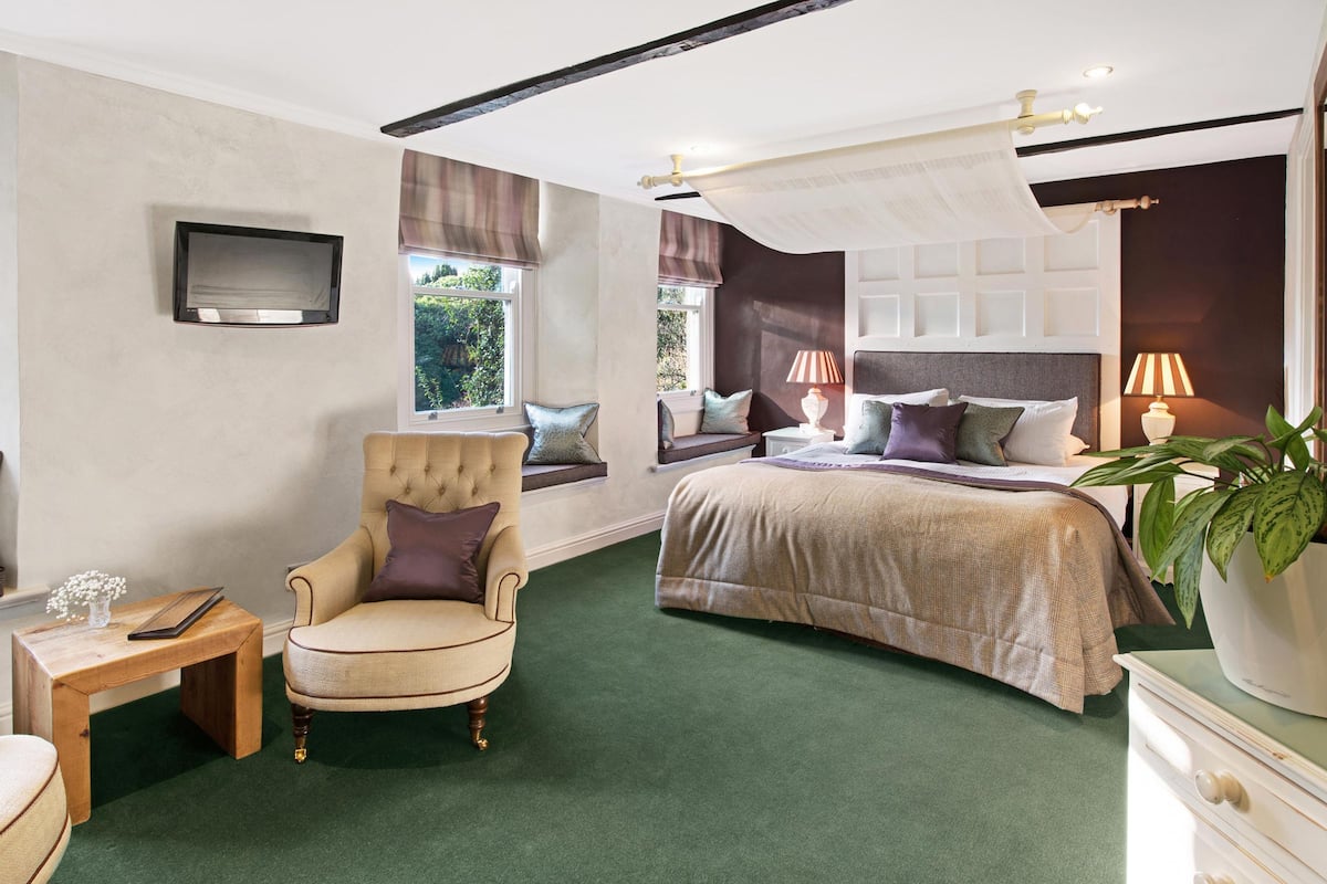 Un dormitorio con cama, una silla, una mesita de noche, televisión y ventana con cortinas.