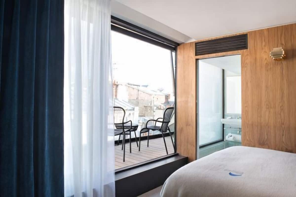 A hotel room with a large window, a balcony with chairs, a wooden wall, and a bed.