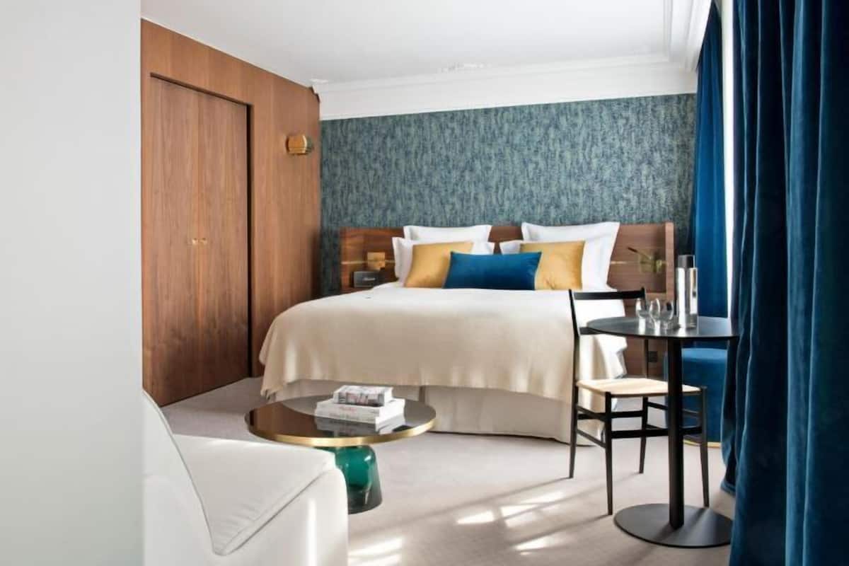 A hotel room with a bed, a round coffee table, a chair, a small table with glasses, and a wooden wardrobe.