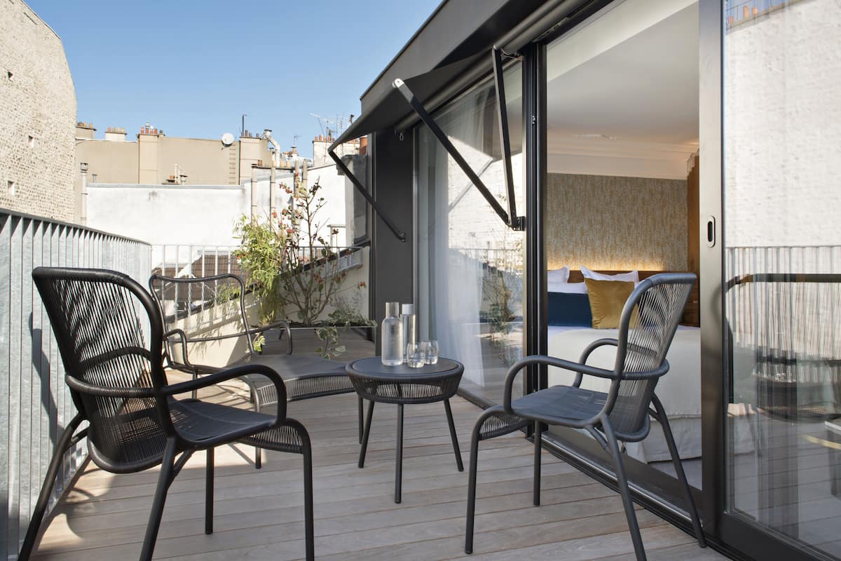 A balcony with two chairs and a small table, a sliding glass door leading to a bedroom, and a view of the cityscape.