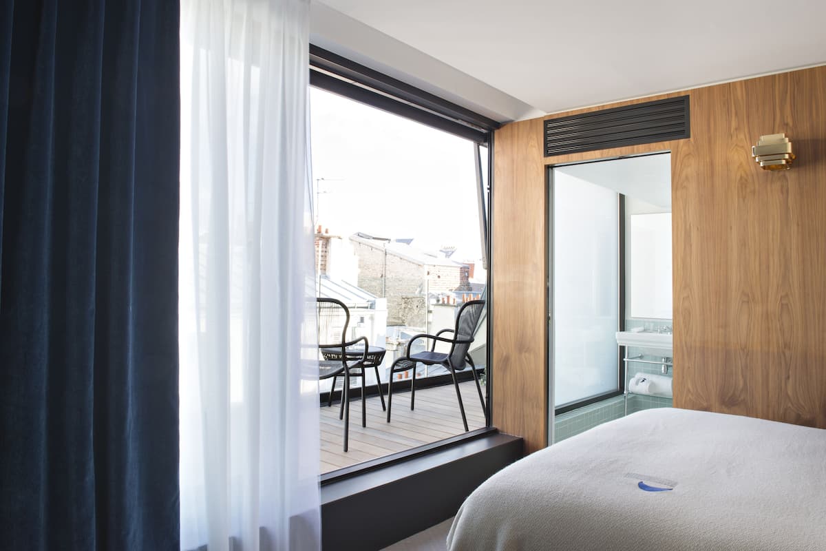 A modern hotel room with a large window, a balcony with chairs, and a wooden wall panel.