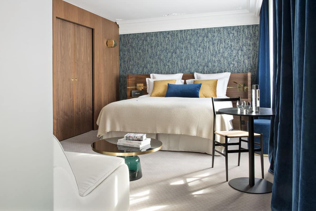 A hotel room with a bed, a round coffee table with books, a chair, a small table with glasses, and a nightstand.