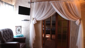 A room with a leather armchair, a flat-screen TV, a fan, and a sliding glass door with white curtains. - Amjicaja Guesthouse (Walvis Bay)