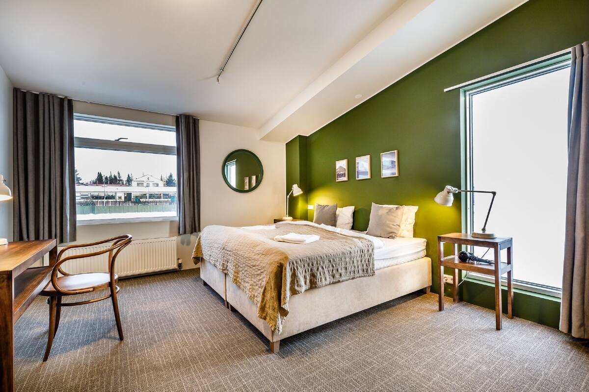 A hotel room with a bed, a desk, a chair, a window with curtains, a round mirror, and a green accent wall.