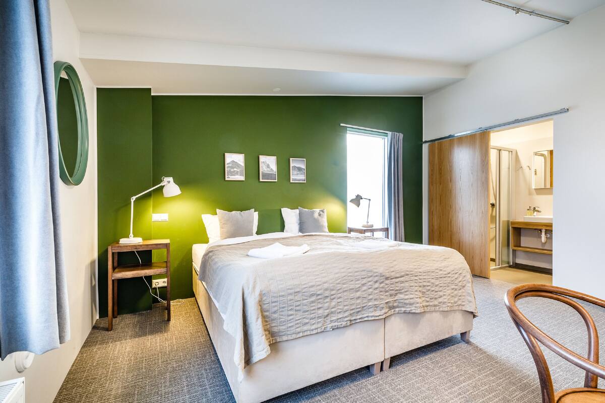 A hotel room with a large bed, a nightstand, a desk, and a chair. There is a green wall, a window, and a door leading to a bathroom.