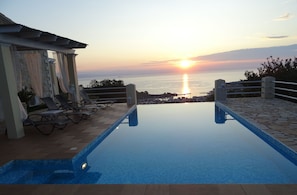 An infinity pool with a view of the sunset over the sea, lounge chairs, and a pergola.